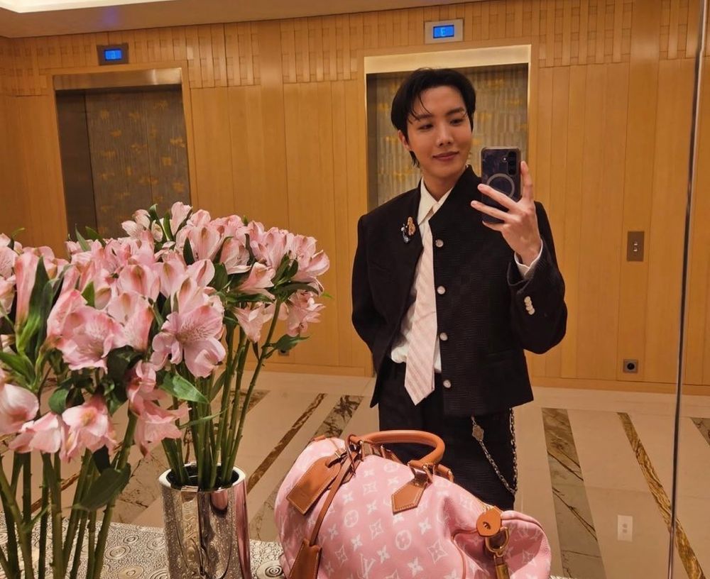 a mirror selfie of hobi in a hotel hallway in front of the elevator bank. he's wearing a Louis Vuitton suit with a light pink tie and matching pink bag, and he's placed the bag on a table next to a vase of flowers in the same shade of pink. he looks very pleased with himself.