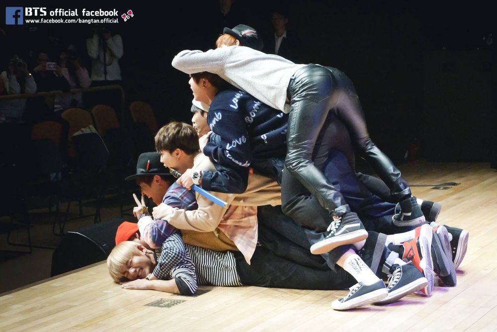 bts ot7 on stage at a 2014 fansign. taehyung is lying face down on the stage with the other 6 piled atop him. the photo is taken from behind and to the side, and yoongi, on top of the pile, is wearing tight leather pants so the overall is effect is of a pile of boys with a very round and shiny ass atop it.