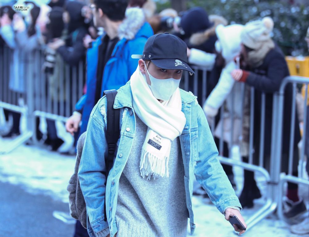 j-hope arriving to the 2016 kbs song festival. he's dressed in a gray sweater, white scarf, jean jacket, and black baseball cap with a skull on the brim. he's wearing a mask and looking up from under the brim of the hat in a brooding and menacing way.