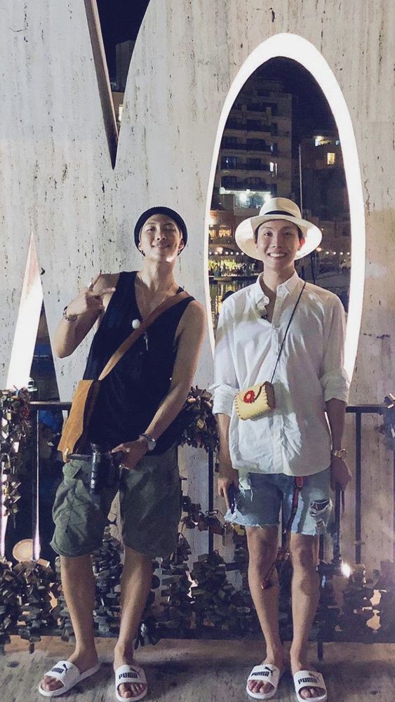 namjoon and hobi in malta, wearing slides and shorts. hobi is wearing a small wicker bag with a flower clasp that namjoon bought for him while filming bon voyage 3.