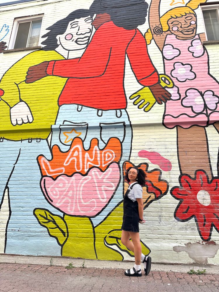 photo of sheila posing in front of a mural of people dancing with the words “land back”