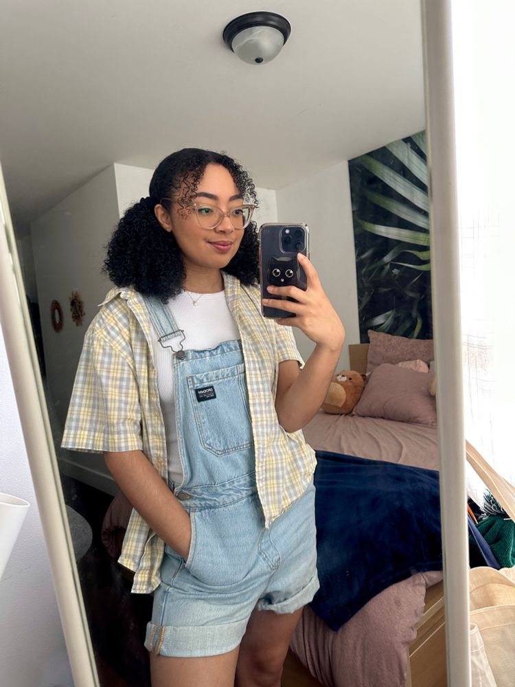 mirror selfie of sheilur wearing a plaid light yellow shirt over light blue overalls, with her bedroom in the background. 