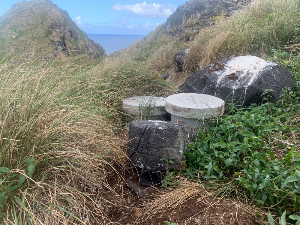 Two circular concrete caps sit atop cylindrical nesting chambers set in grass tussocks, with weighty rock semi/collapsing a tunnel entrance in the foreground. Background shows two peaks on a steep slope with ocean behind.