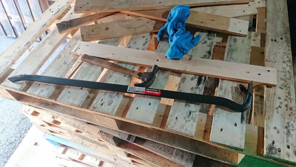 A long crowbar atop a pile of pallets with some separated planks on top too; the crowbar is almost as long as the pallet is wide, somewhere around 3ft/1m long.