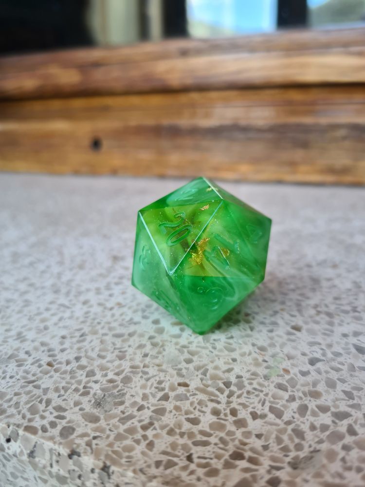 Big d20 on a flat surface. It is green with white swirls and small pieces of gold leafs.