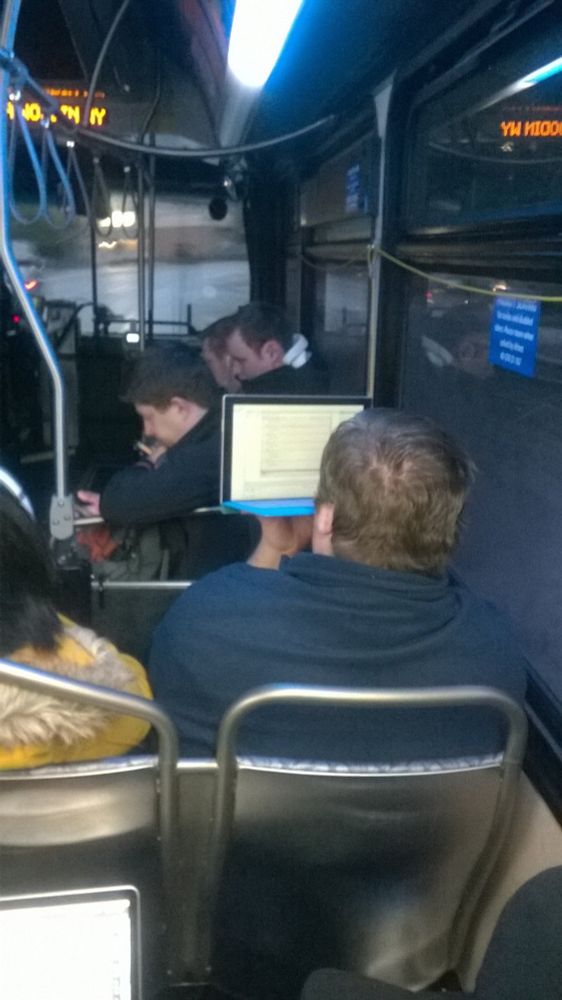 Man riding the bus holding his laptop up in the air to work.
