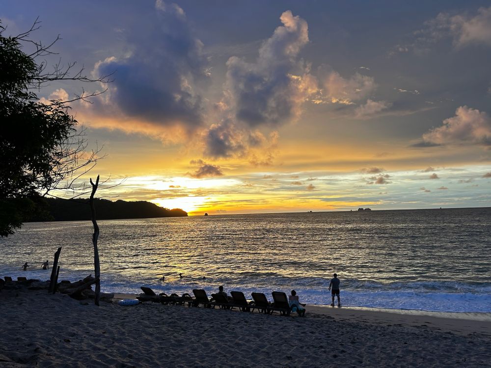 A view from a beach out onto the Pacific Ocean as the sun sets. A line of chairs and a lone silhouette of a figure are on the beach as a wave crashes. The sunset is shades of yellow and orange on the distant horizon. Layers of clouds drift on by.
