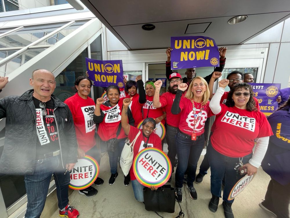 People in red tshirts posing at an airport picket with UNITE HERE and UNION NOW signs.