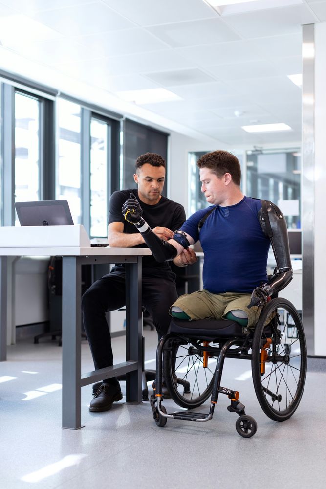 A man in a blue shirt and khaki shorts sits in a wheelchair (his legs are amputated at the knee), getting his prosthetic right arm adjusted by a technician in a black t-shirt inside a brightly lit office suite. The prosthetic on the man's left arm is appears suitable attached.

📸: ThisIsEngineering