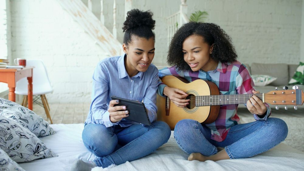 Color photo: two cheerful teenage girls sit atop a made bed. One is playing a guitar while the other is holding a phone for the guitar player so she can see the lyrics/chord changes.

📸: Vitaly Gariev