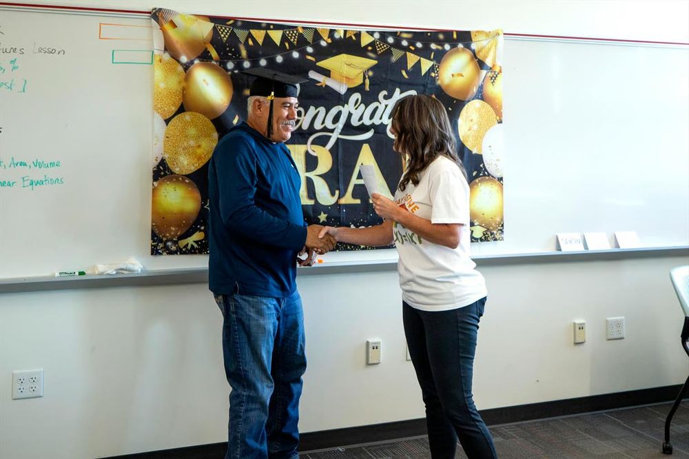 A man and woman shake hands in front of a congratulatory banner of gold balloons on a black background and the phrase "Congratulations GRADS" pinned to a wall-length whiteboard. The man has a grey mustache and wears a tasseled graduation cap. The woman's face is hidden by her hair.

📸: Kevin J. Beaty/Denverite