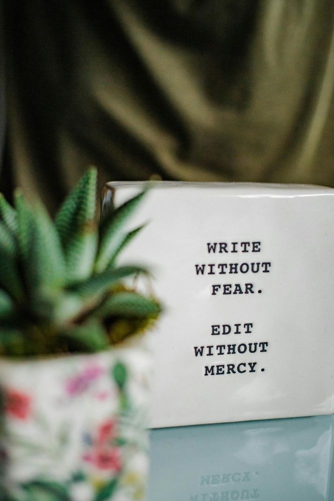 A small white plaque with the phrase "Write without fear. Edit without mercy" sits on a glassy tabletop. A potted succulent in a floral pot is slightly  out of focus in the foreground. Rippling green fabric fills the background.

📸: Hanna Grace