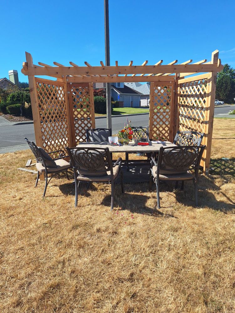 A cedarhrape arbor structure near the street with a table and chairs placed underneath it, on a golden dead lawn