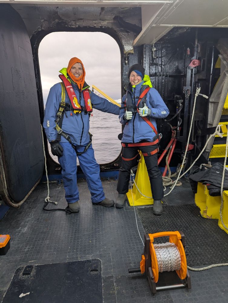 Thom Linley and Kat Bolstad stand in front of an open hatch in safety gear. The ocean and icebergs are visible through it. 