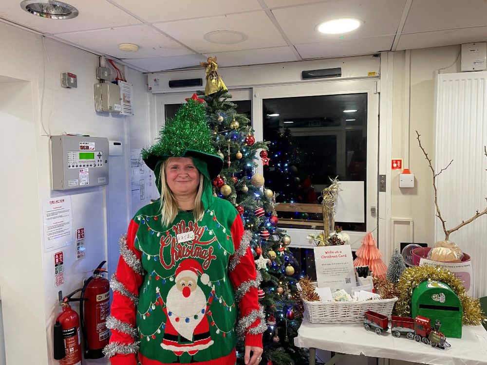 Kath from GL11 wearing a green Christmas jumper with Santa and a matching green glittery hat standing next to a decorated Christmas tree indoors.