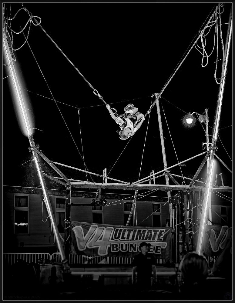 Black and white night photo of a young boy attached to bungee cords around his waist in a large V shaped frame, with a trampet on the floor beneath him; caught mid-somersault 20 ft in the air