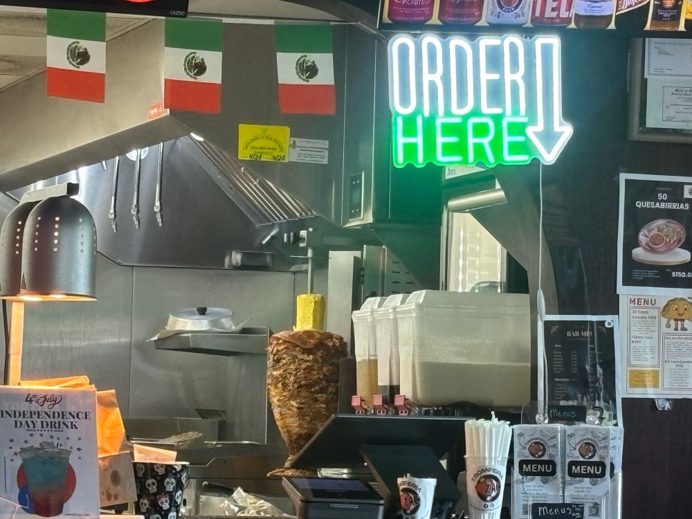 The counter at Tromperia el Zac in Saint Paul - a white and green sign reading “Order Here” and a trompo (meat cone) behind the counter, ready for cutting.