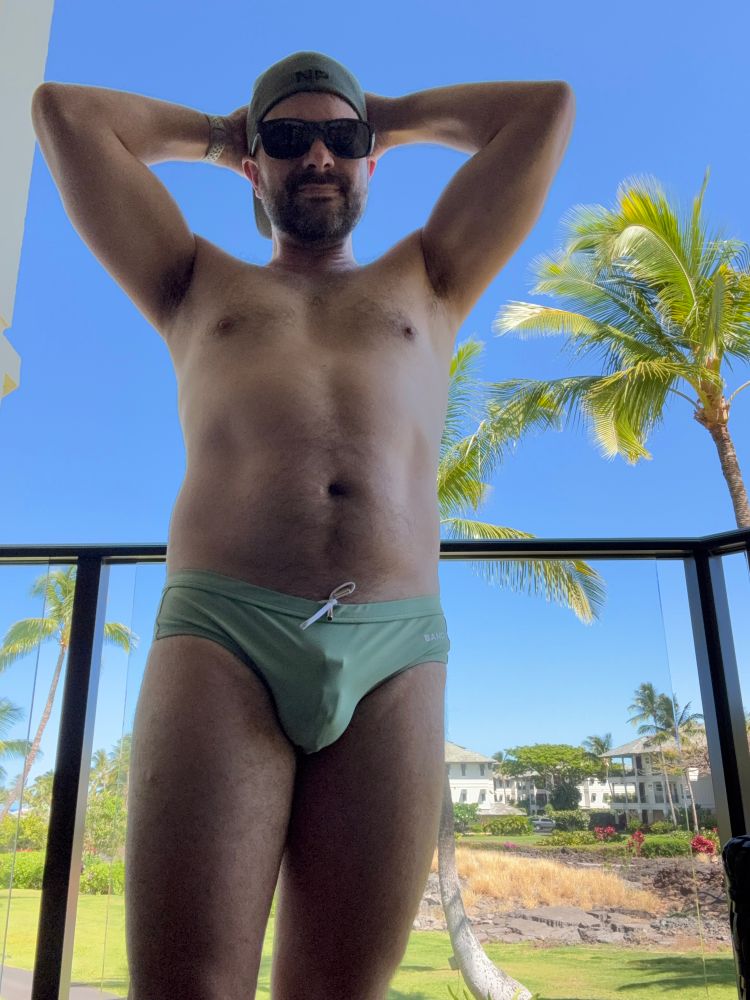 A man standing on a balcony wearing a green speedo. The bulge from his chastity cage is visible.