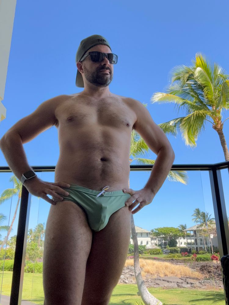 A man with his hands on his hips standing on a balcony wearing a green speedo. The bulge from his chastity cage is visible.