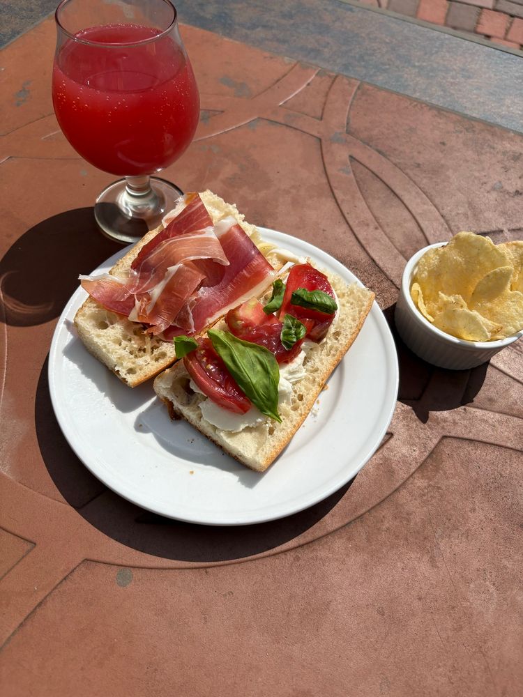 Sandwich of butter, prosciutto, mozzarella, and basil on a baguette, side of chips, glass of San Pellegrino blood orange with a splash of lemonade, on the back yard patio table