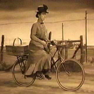 yellow toned photo still from the Wizard of Oz, showing the character Miss Gulch biking down the road with a basket behind her on the bike