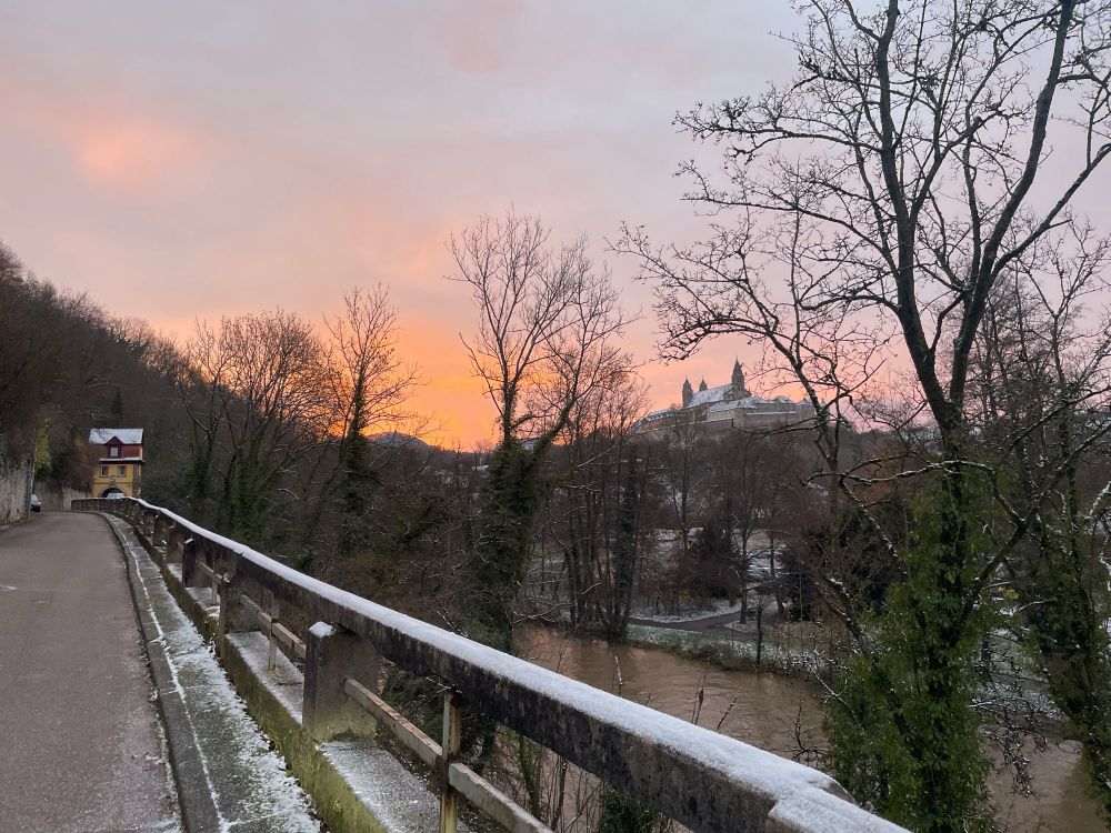 Morgenrot über einem alten Kloster auf einer Anhöhe. Im Vordergrund sieht man Bäume, einen Fluss und ein schneebedecktes Gelände aus Beton.