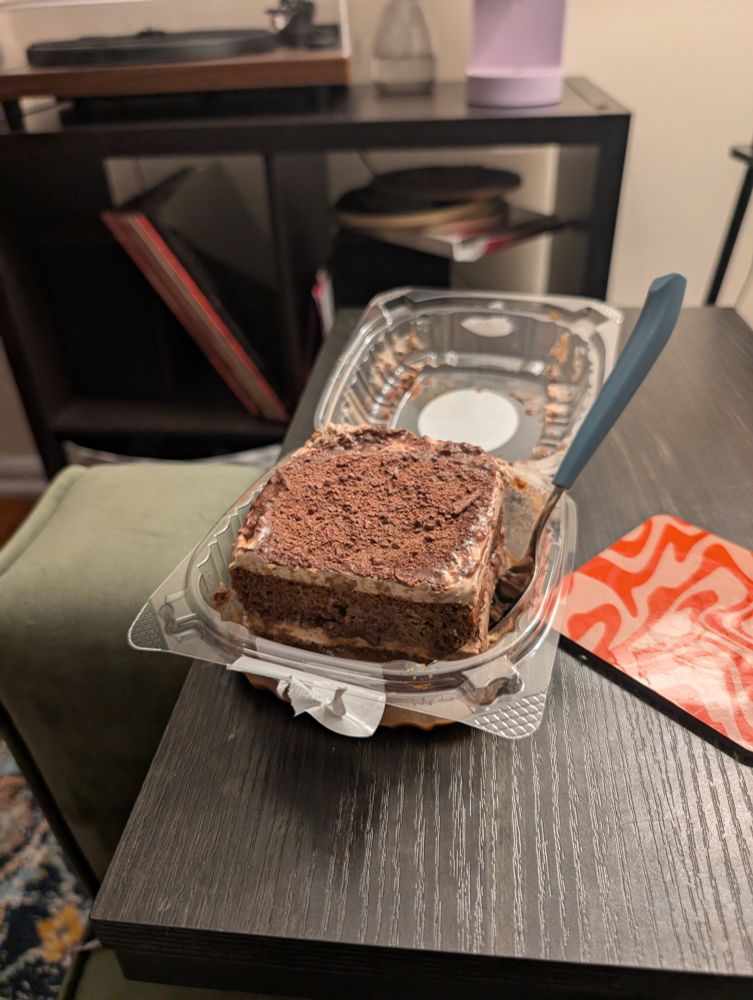a plastic container filled with a large square or tiramisu with a spoon sticking out of it I'm going to attempt to eat the whole thing 