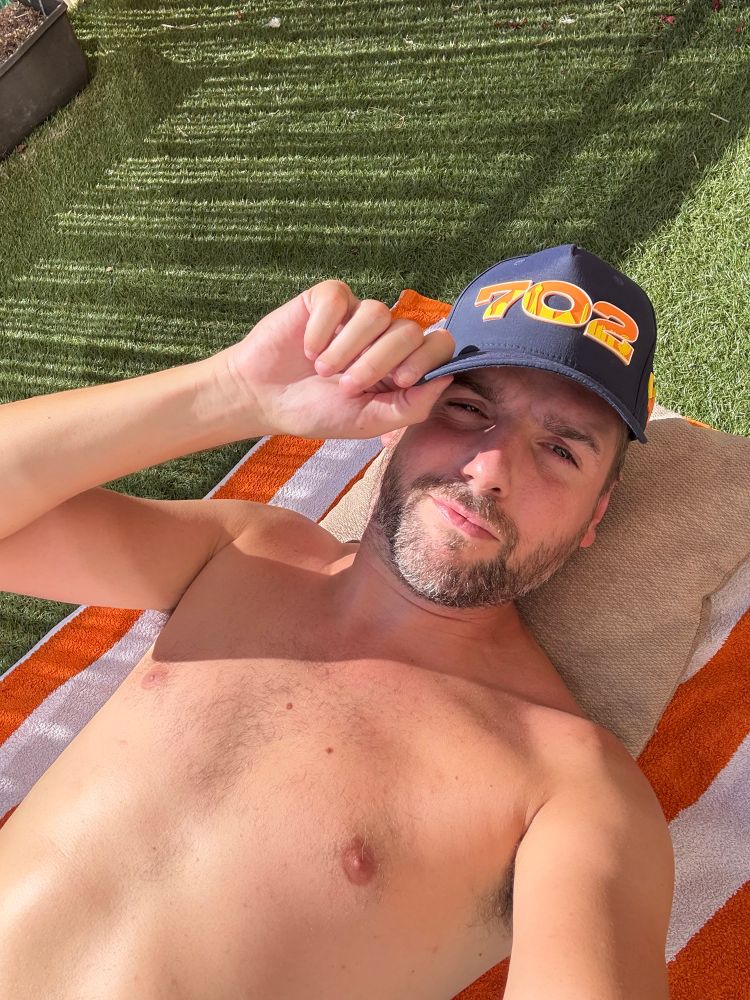 Matt laying on his terrace in the sun, tanning and wearing a navy baseball cap with the numbers 702 printed on the front. 