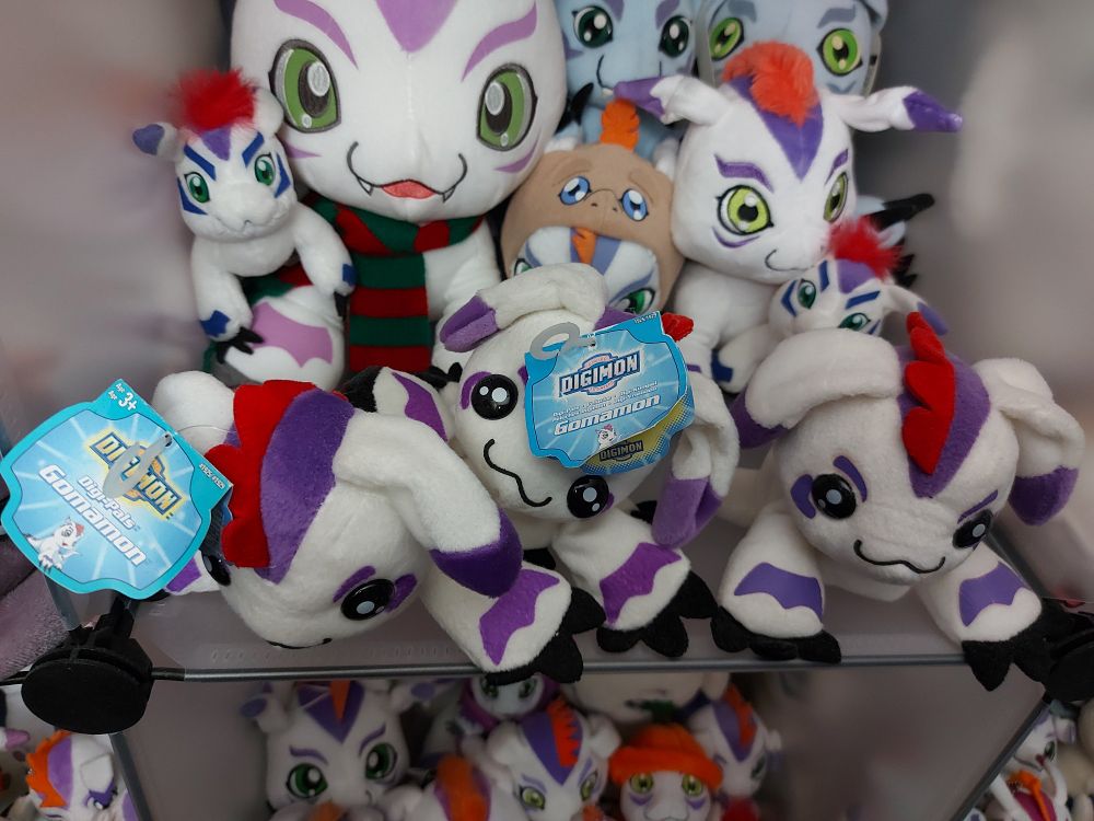 A photo of digimon (Gomamon) plushies in a modular cube shelf. The 3 in the front are similar but with differences in tag text or markings print depending on where they were made. (Left: North America, centre: Europe, right: Japan)