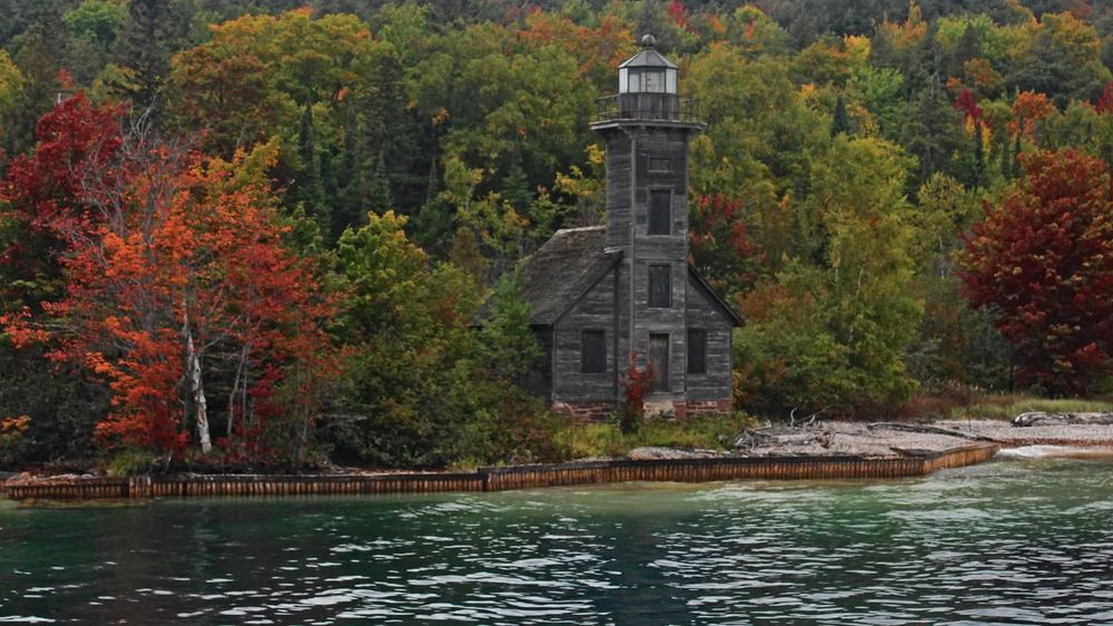 A dark, rickety wooden lighthouse on the shore of a dark green watered lake. Behind it is a field of green trees tinged yellow, and on either side are two orange trees turned a brilliant orange. 