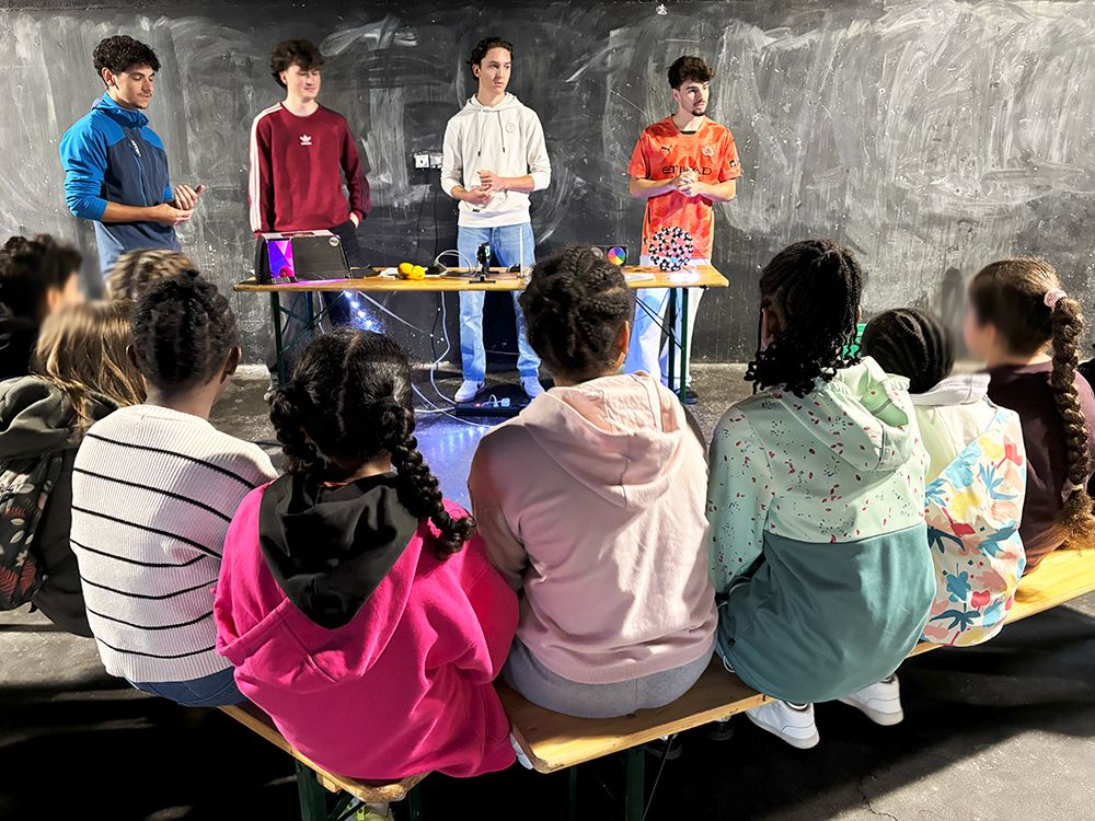 4 étudiants présentent des expériences scientifiques. Ils sont debout, devant une table. Au premier blanc, plusieurs enfants (de dos) sont assis sur des bancs et écoutent les étudiants.