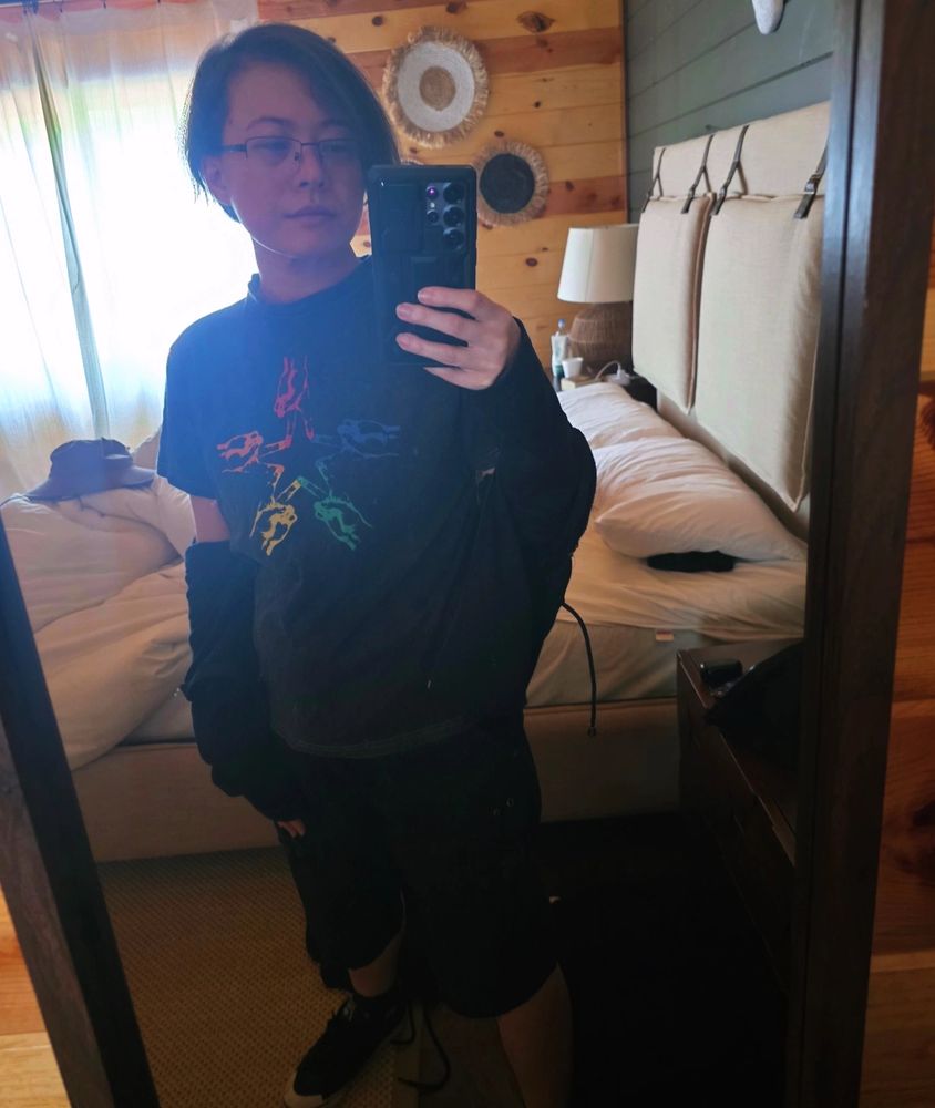 OP taking a mirror selfie far from home. he's got a shirt with rainbow hands in the shape of a star on it.