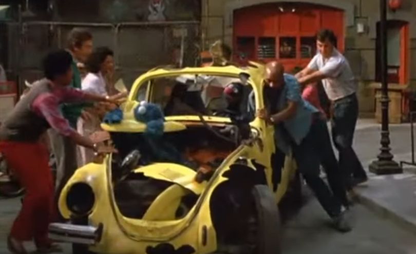 a yellow vehicle, driven by Grover, full of holes being pulled into Sesame Street by many humans. Cookie Monster, displaying biblical greed, is sitting where the engine should be and eating the vehicle's hood.