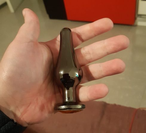 In meiner weißen Hand ist ein metallener, mittelgroßer Buttplug mit einem orangenen Stein am Ende.
Im Hintergrund ist ein roter Teppich am unteren Bildrand zu sehen, darüber graußer PVC Boden und eine rote Schranktür.