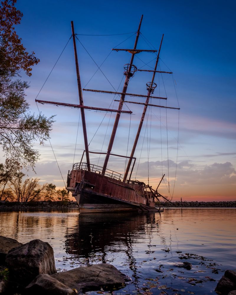 A rusty old beached ship with three masts photographed at sunset with rocks in the foreground and trees along the left side. The sky is gold and blue with low clouds along the horizon. The sky’s colours are reflected in the still water