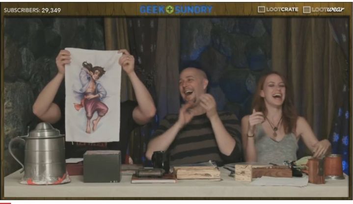a screenshot from Campaign 1 of Critical Role. Sam Riegel holds up a small body pillow of his gnome bard Scanlan Shorthalt to the screen, while fellow players Taliesin Jaffe and Marisha Ray laugh.