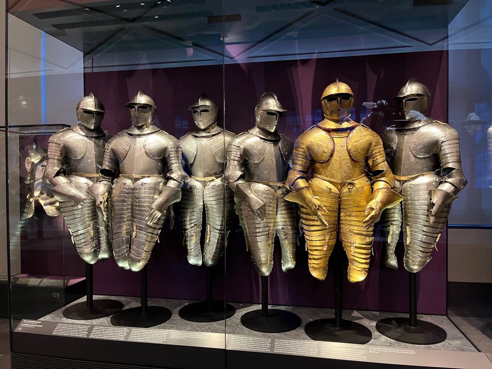 This image shows the full armour flanked by two of the etched silver metal suits of armour. 

Each includes a fitted helmet with openings for eyes, nose and mouth (called a Savoyard burgonet), the metal gorget worn over the armour to protect the throat and upper chest, the articulared pauldrons to cover the shoulders, forearm covers and gauntlets, and knee-length leg armour (called tassets).
