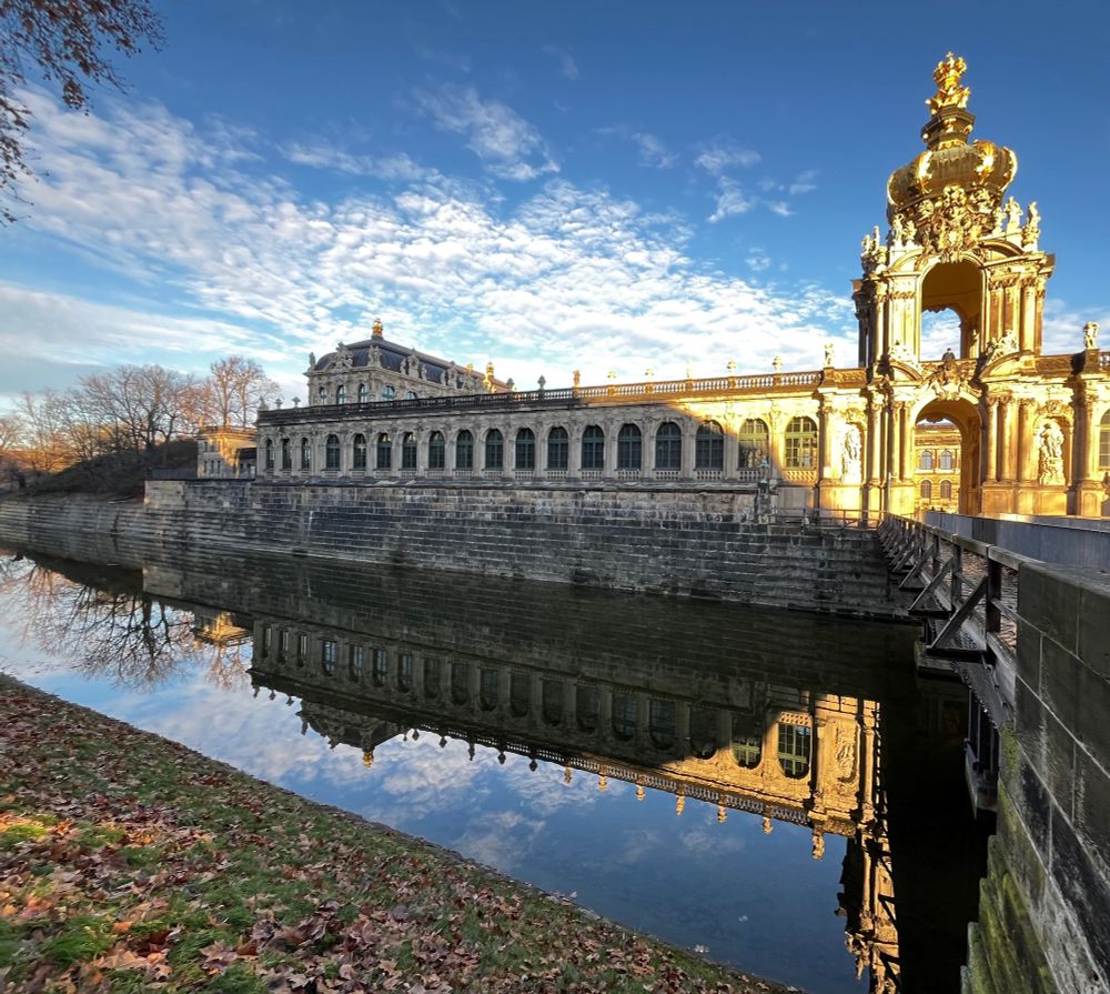 The Dresdner Zwinger, in Dresden, Germany is a Baroque-style grand palace.  Today, it houses world-class museums and an Old Masters gallery.

Partially surrounded by a water feature, this view looks out from the beginning of the footbridge across the water to the two-level, open gateway (on the very right of the image) that stands at the halfway point of the length of the building. The corner of the building we see is half of one of the long galleries. It stretches out to the left, with 18 arched windows, each with 18 panes. Above the row of windows there is a balustrade at the roofline along the rooftop walkway, punctuated with statuary above every space between the windows below.

Captured on a blue-sky/white clouds December day, this wing of the Zwinger is almost totally in shade from the Dresden State theatre across the street (not in the image).  However, the gateway glows in sunshine, partly because of the gilt on the huge crown above the open archway.

In the water feature below, there is a mirror-like reflection of the building, as well as of the leaf-less winter trees at the extreme left of the image. In the immediate foreground there is the leaf-strewn, steep grassy bank down to the moat, leading away from the viewer’s position.

https://www.der-dresdner-zwinger.de/en/the-dresden-zwinger/ 

