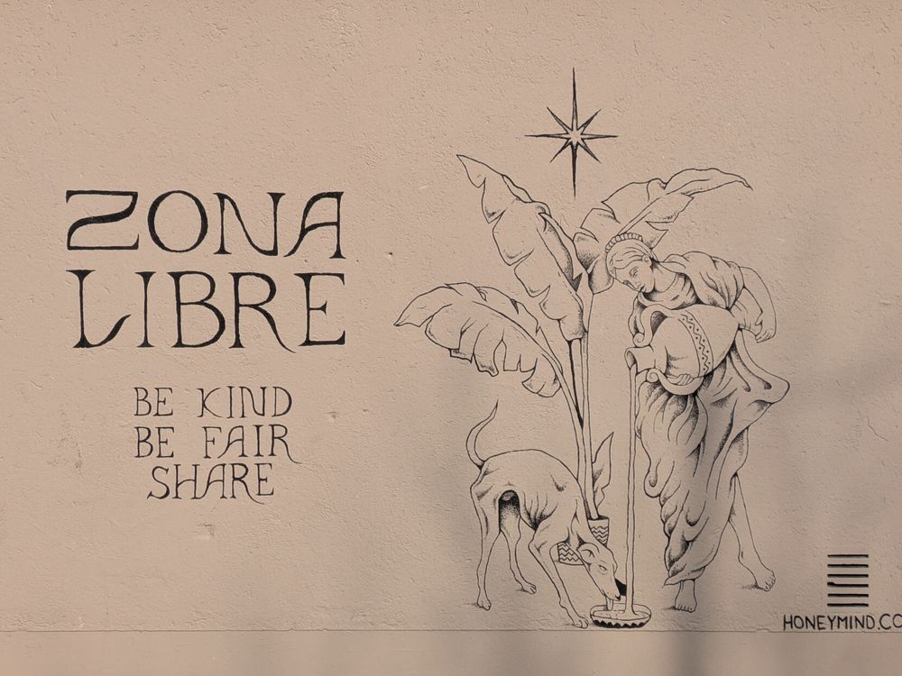 Hellbeiger Putz mit schwarzer Pinselzeichnung: 
Die linke Hälfte in künstlerisch gestaltetem Font 
ZONA LIBRE
BE KIND
BDE FAIR
SHARE
Die rechte Hälfte eine von einem 8-zackigen Stern gekrönte großblättrige Staudein einem Topf, links davon ein sehr schlanker Hund, der aus einem Gefäß trinkt, in das in einem langen Strahl von oben Wasser aus einem Amphore geschüttet wird. Das macht einen antik anmutende Frauengestalt, barfüßig in wallendem Gewand.
Rechts unten der Name HONEYMIND.COM
Das Wandbild wirkt wie ein Kunstdruck.
