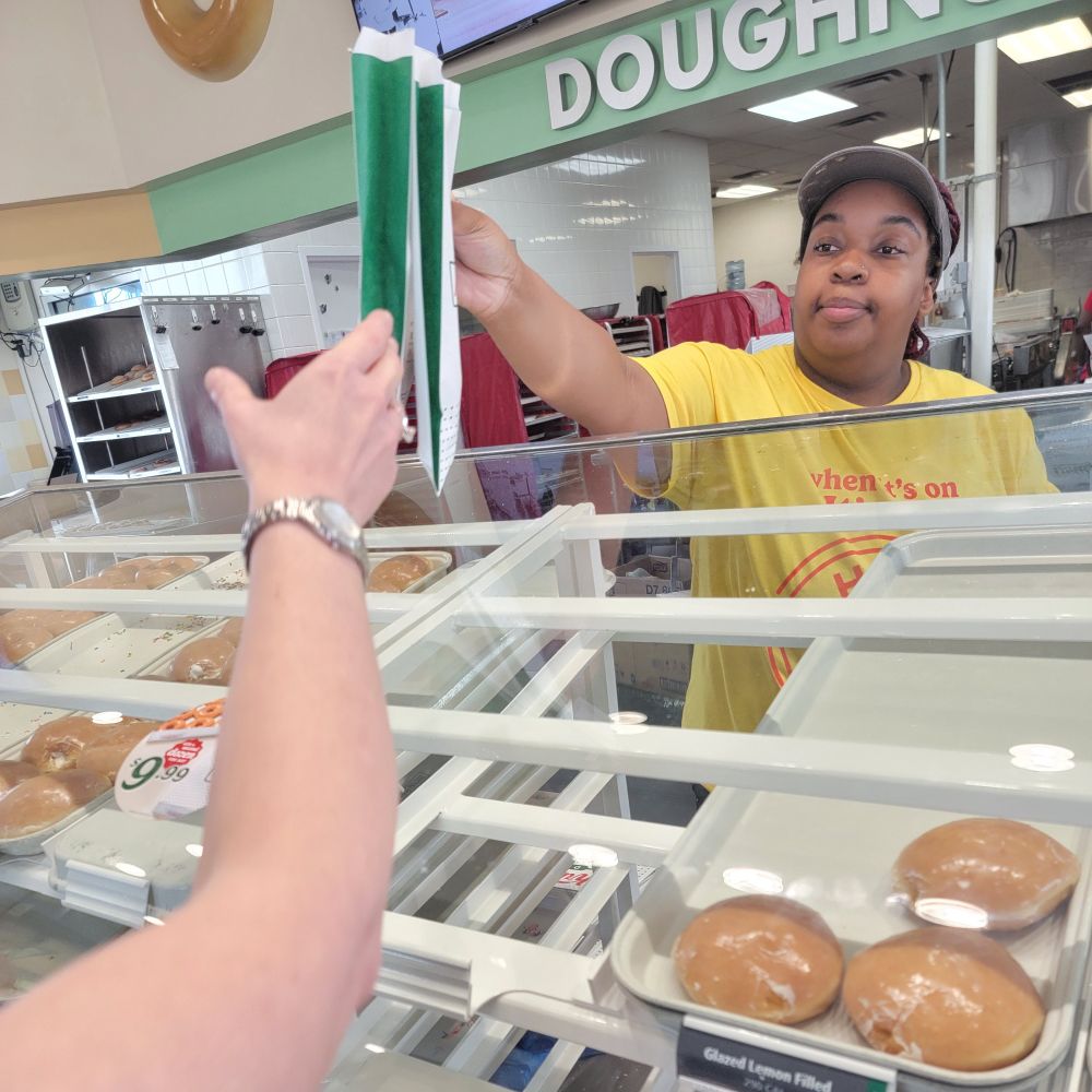 A Krispy Kreme employee behind the display counter hands two bags with doughnuts to the outstretched arm of a customer.