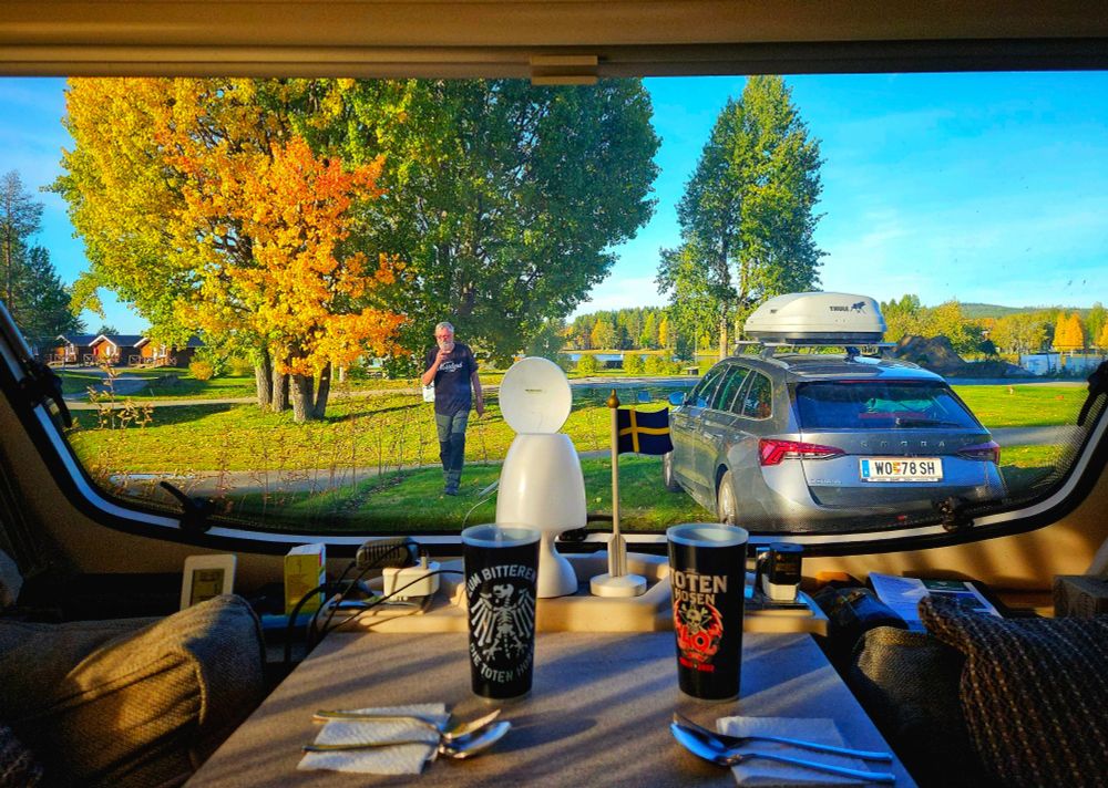 Blick aus einem Wohnwagenfenster auf einen herbstlichen Campingplatz: Im Vordergrund ein gedeckter Tisch mit zwei Bechern, Besteck und einer kleinen schwedischen Fahne. Draußen steht ein graues Auto mit Dachbox neben einem großen Baum mit grün-gelben Blättern, teilweise leuchtend orange gefärbt. Ein Mann geht auf den Wohnwagen zu, im Hintergrund sind weitere Bäume, Rasenflächen und kleine Holzhäuser zu sehen.
