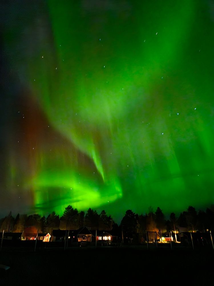 Ein riesiges spiralförmiges Polarlicht in Grün- und Rottönen strahlt am Himmel. Am unteren Bildrand sind beleuchtete Häuser zu erkennen.