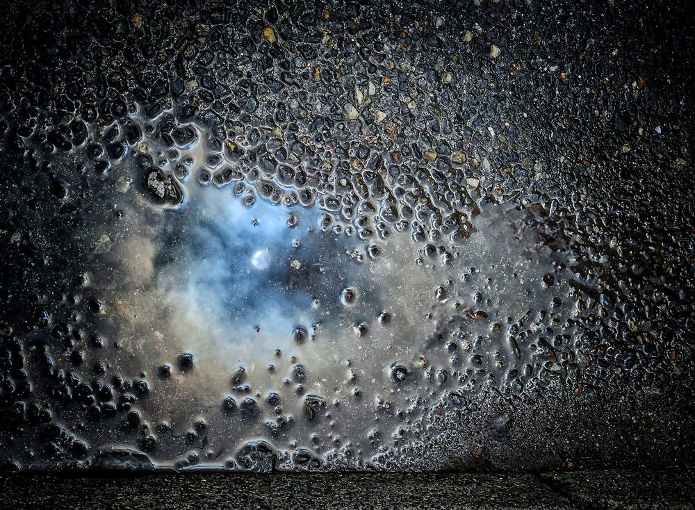 Blick von oben auf eine grob asphaltierte Straße mit einer kleinen Pfütze. Die Sonne und abziehenden Regenwolken spiegeln sich im Wasser, sie sehen aus wie ein Mini- Universum. Am unteren Bildrand ist eine Bordsteinkante zu erkennen.