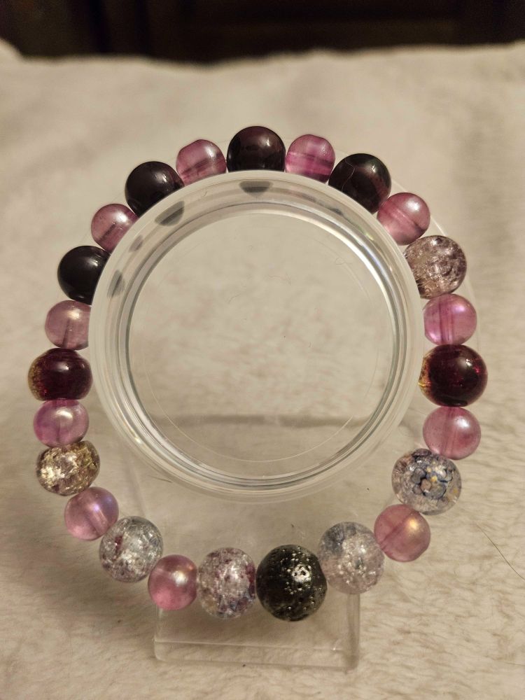 Handmade glass bead bracelet on a clear display stand and a light tan background. Bracelet is a mix of solid dark purple, crackle purple/yellow, crackle white, crackle pink, and smaller clear bubble pink beads in a gradient from light at the bottom to dark at the top. It features a bright metallic black colored lava volcanic natural stone bead at the bottom