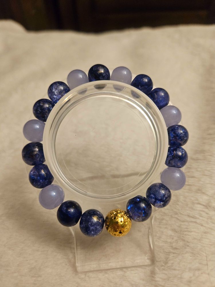 Handmade glass bead bracelet on a clear display stand and a light tan background. Bracelet is a mix of crackle blue, crackle dark navy blue, light grey blue beads. It features a bright metallic gold colored lava volcanic natural stone bead at the bottom
