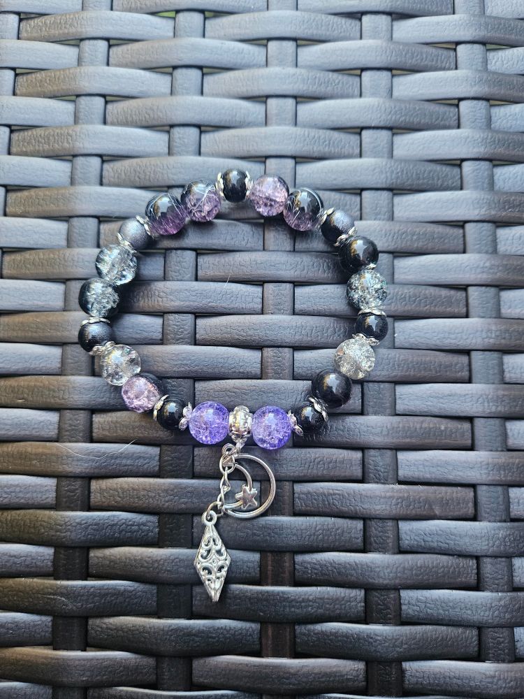Handmade glass bead bracelet for goddess Hekate. Bracelet is displayed on a dark brown basket weave pattern background and features two silver charms: a moon outline with a star attached and a filigree diamond shape on a silver extender chain. Beads are crackle purple, solid black with half silver, silver crackle, black/purple crackle. Silver bead caps accent the beads