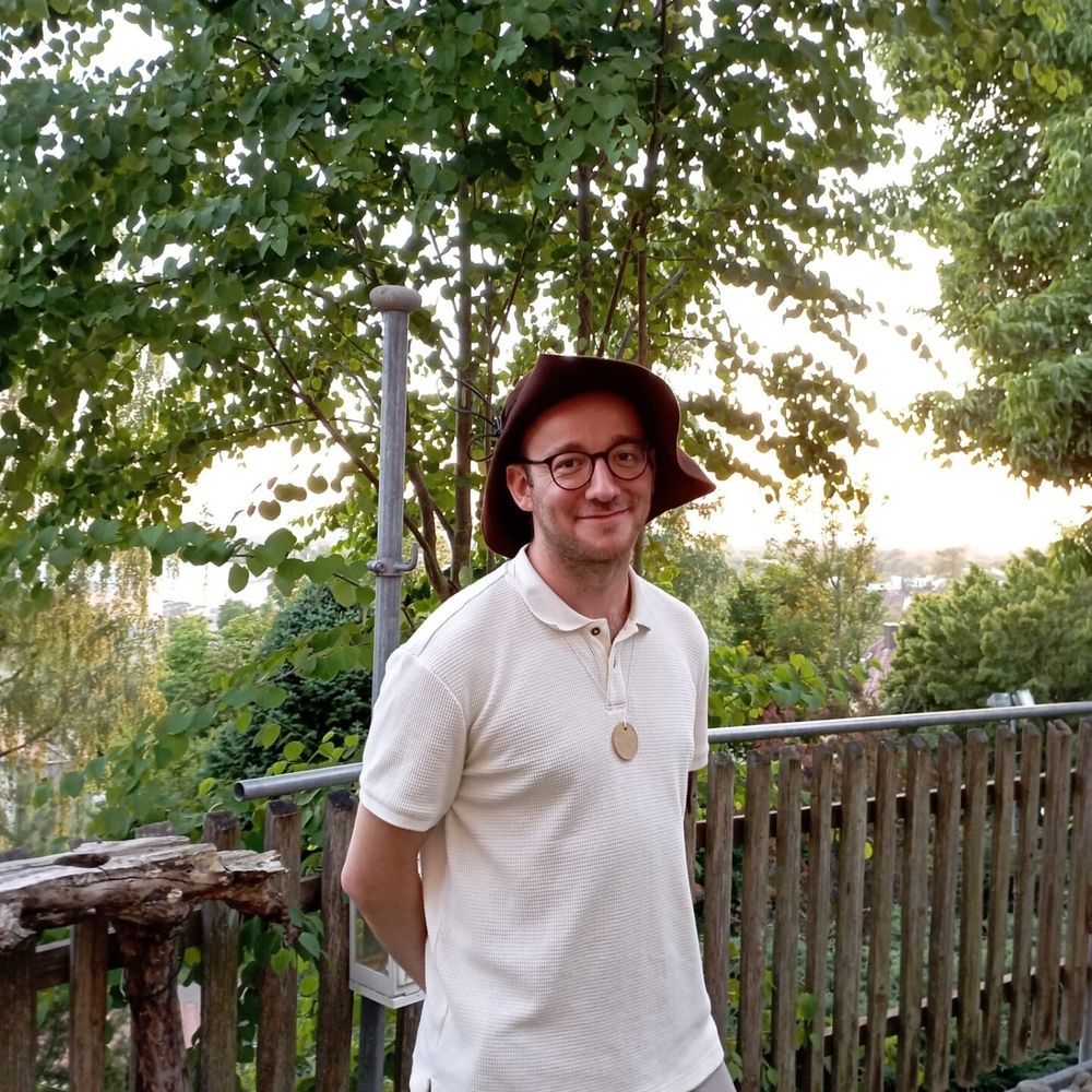 Dax Werner steht lächelnd vor einem hölzernen Zaun im Grünen. Er trägt ein beiges Poloshirt mit einem runden Holzanhänger und hat die Hände hinter dem Rücken verschränkt. Im Hintergrund sind Bäume und eine leicht hügelige Landschaft bei Sonnenuntergang zu sehen.

