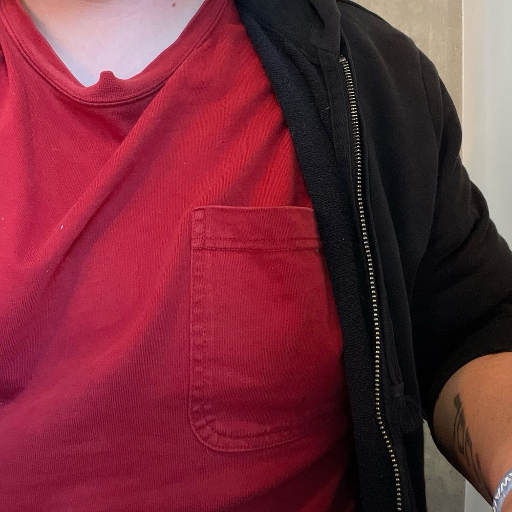 Photo of the chest of a white butch person wearing a red pocket tee and black hoodie 