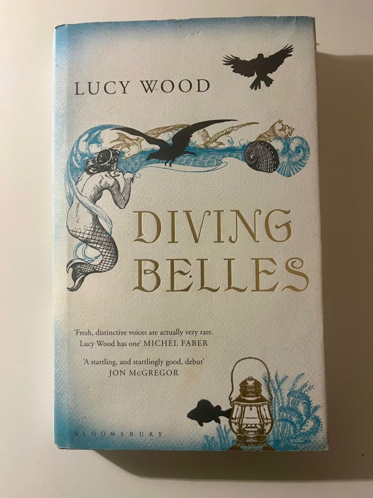 Hardback Bloomsbury edition of ‘Diving Belles’ by Lucy Wood. The front cover features a surfacing mermaid peering at a twist of blue and gold waves, gulls and shells. In the bottom right corner, a fish swims by a sunken lantern. 