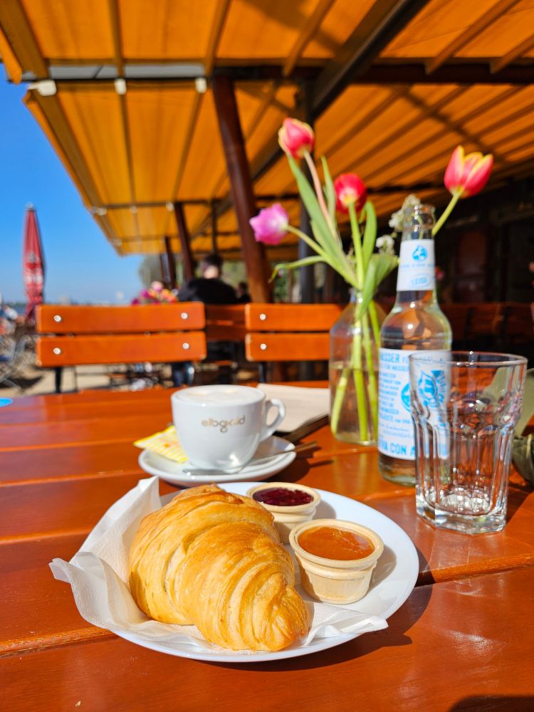 Croissant, Marmelade, eine Tasse Kaffee, eine Flasche Wasser, im Hintergrund eine Vase Tulpen auf einem Holztisch. Ein bisschen blauer Himmel ist zu sehen.
